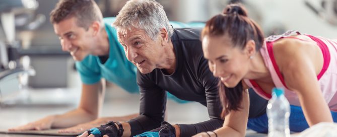Group of people in various age and gender having fun while doing group exercise of elbow plank in the gym.
