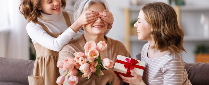 Happy International Mother's Day.Smiling daughter and granddaughter giving flowers and gift to grandmother celebrate spring holiday Women's Day at home