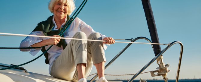 Enjoying sea trip. Happy senior woman sitting on the side of a sail boat or yacht deck floating in sea, looking at camera and smiling. Clear blue sky background